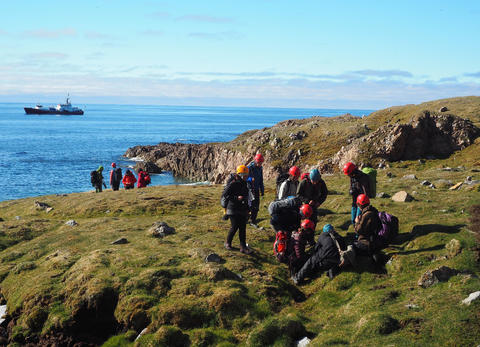 Students surveying the grassy vegetation on the coast of Svalbard with a ship on the sea in the background