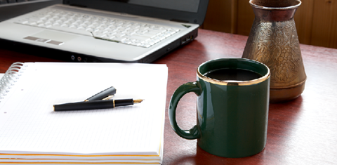 Bilde av skriveblokk med penn og kaffekopp med laptoppen i bakgrunnen