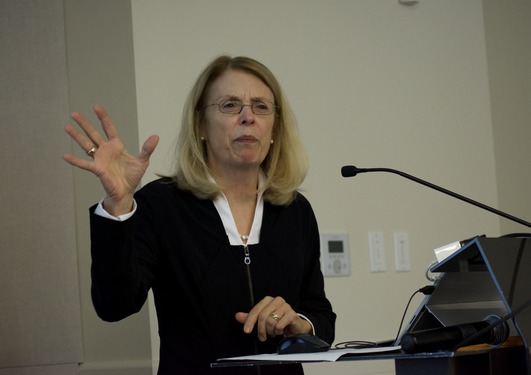 A blonde woman in black top and glasses is speaking in a microphone, holdling one hand up in the air