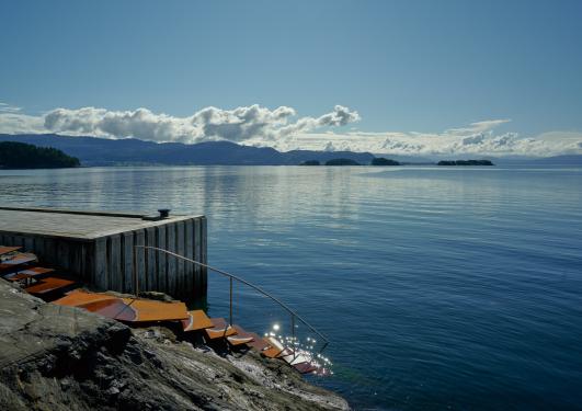 Bildet viser Bjørnefjorden med utsikt mot andre siden. Det er en trapp ned i fjorden og en brygge ved siden av trappen. Fjorden og himmelen er blå og imellom er lette skyer i horisonten.