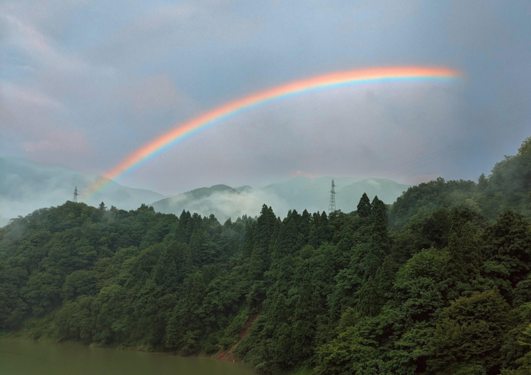 Landscape with a waterbody in front, surrounded by thick, dark green forest on the right. Behind  the forest peaks mist-covered mountains. Half a rainbow is visible across the sky, framing the mountain tops.