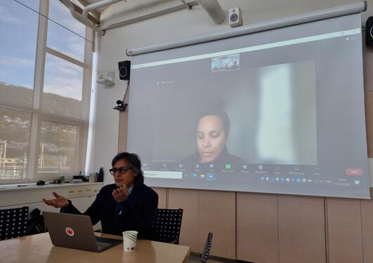 A person is sitting at a table in front of a computer, behind them is a person participation in a live Zoom call