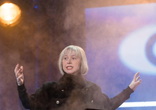 Kvinne på scenen, omringet av røyk og dyster stemning. Vi skimter et øye i bakgrunnen.