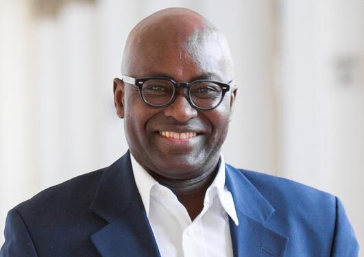 Profile picture of Achille Mbembe. He is a black man with glasses, wearing a blue suit with a white shirt and smiling. 
