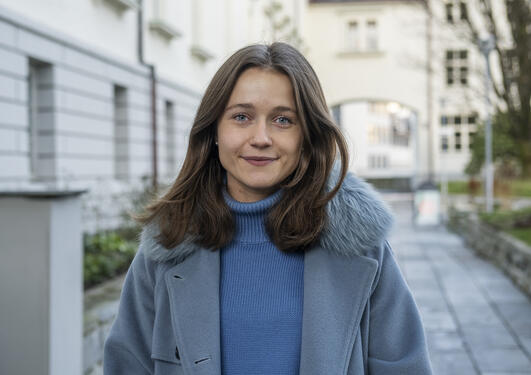 Anne-Line står utenfor universitetet med en gul/hvit bygning i bakgrunnen. Blått kaldt lys.