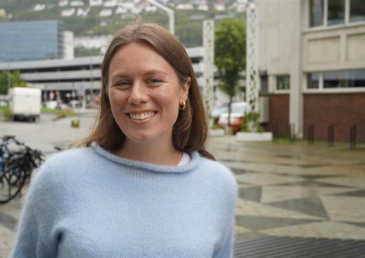 Kvinne med brunt hår som smiler. Bygarasjen kan ses i bakgrunnen. 