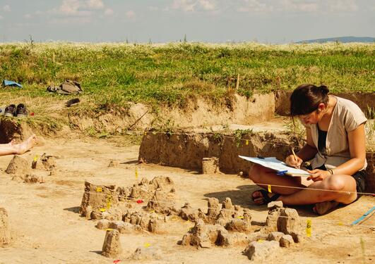 Woman sitting tailoring position on the ground making notes in the middle of an excavation.