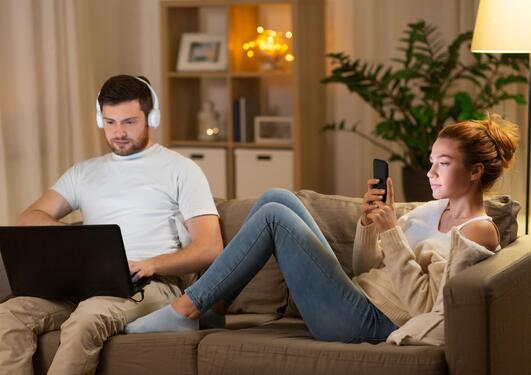 Par som sitter i sofaen på kvelden med laptop, hodetelefoner og smarttelefon