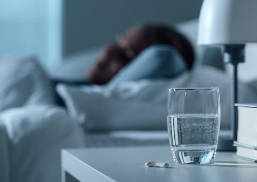 glass med vann og medisiner i forgrunnen - i bakgrunnen, ute av fokus, en kvinne som ligger på en seng og sover eller hviler 