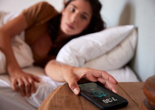 En ungdom strekker seg etter telefonen i sengen, klokken på alarmen viser 07.30