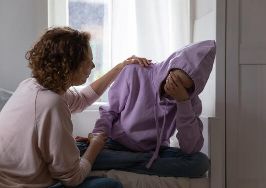 Tenåring med fiolett hettejakke stitter på sengen og blir trøstet av sin mor som holder henne i skulderen og på armen. Tenåringen har hånden foran ansiktet