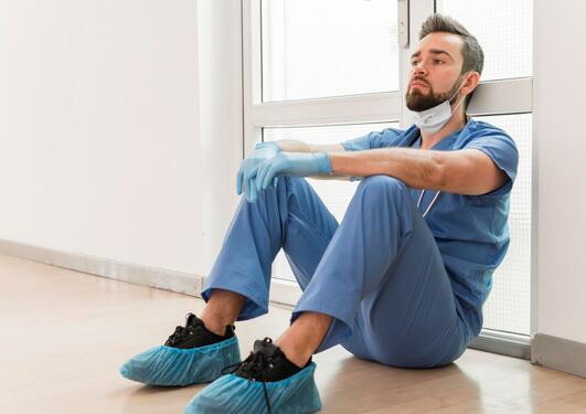 Mannlig sykepleier i blå uniform sitter på gulvet og lener seg mot veggen tydelig sliten