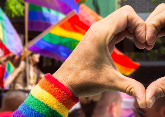 Bilde fra pride parade der noen holder fingrene formet som et hjerte og det er mange regnbueflagg