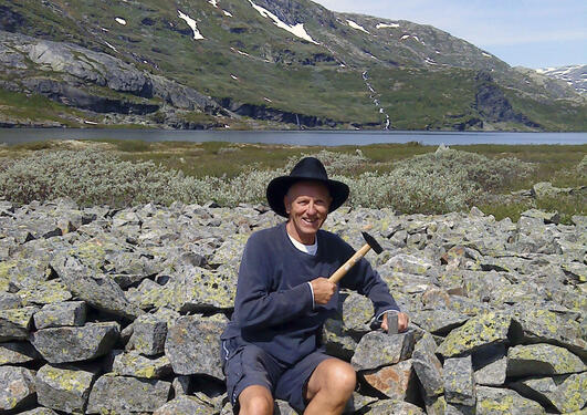 Inge Olav Aarseth sitter med hammer og Rallarhatt oppe i fjellet med et godt smil