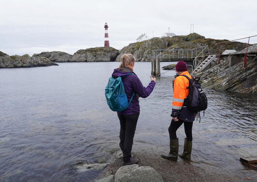 To turkledde personer står i vannkanten med ryggen til, og ser utover fjorden. Fyrtårn i bakgrunnen.