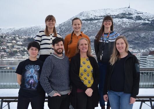 Group of seven people standing outside, smiling. Snow-covered mountain in background.