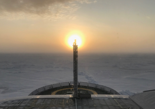 View from the research vessel Kronprins Haakon, overlooking a white sheet of ice with the sun just over the horizon in the center of the photo.