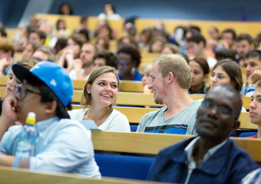 Auditorium med mange studenter og to som ser smilende på hverandre i fokus