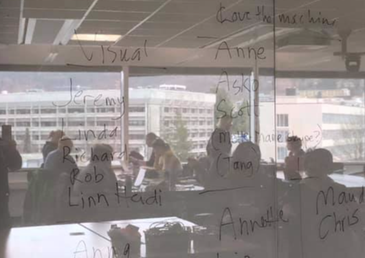 Photo of a wall with names written on it. In the shiny surface of the wall you can see the reflection of people sitting at tables, and a view of buildings seen out of large windows. 