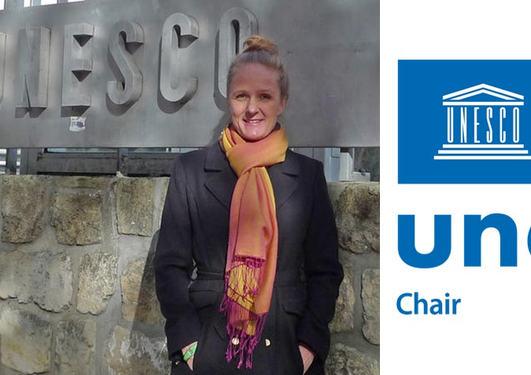 Woman wearing a coat and scarf, smiling in front of large unesco sign. Picture marked with UNESCO chair logo.