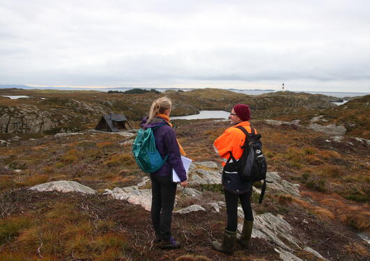 A man and a woman standing in a coastal landscape, talking and enjoying the view. A small cabin in the background, and a fjord and a lighthouse in the horizon. Grey, cloudy skies.