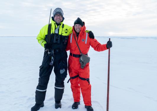 To menn i kjeledress på isen. Alt er hvitt. De smiler til kamera.