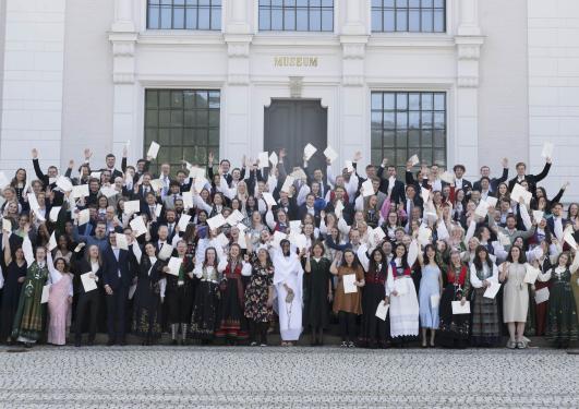En stor gruppe mennesker i bunad og dress står samlet i trappen forran Naturhistorisk museum. Alle er glade og holder diplomet i hånden i været mens de smiler og ler. Dekanen står fremst i midten av gruppen i dekankappe.