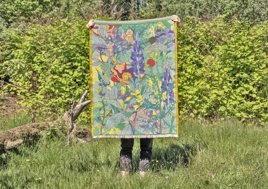 Person holding a colorful, floral-patterned artwork in a lush green garden setting. The person's face is not visible.