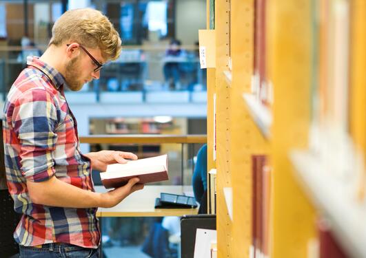 Mannlig student som står og leser ved en bokhylle på biblioteket