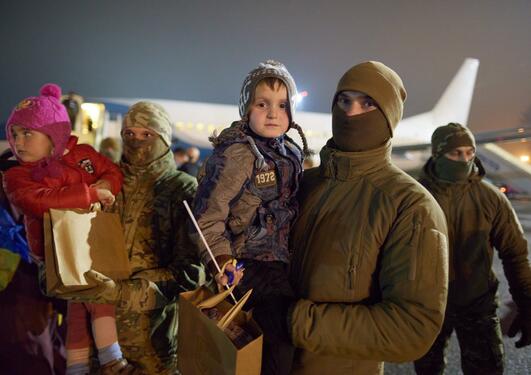 Three men and three children in front of a plane at night time - the man in front has a facecovering and is holding a child