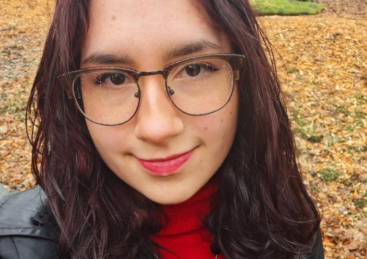 Portrettfoto av person med mørk hår til over skuldra, briller og svart jakke over raud gensar