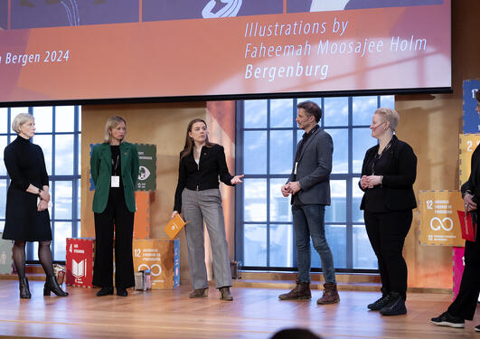 Fice people on the stage engaged in conversation. They are in the university aula and behind them are SDG figures