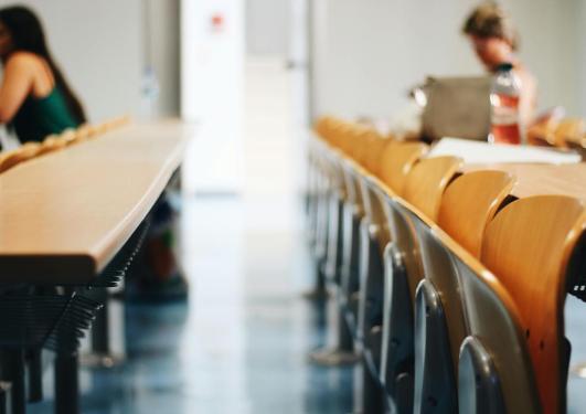 Bildet viser stoler fra et auditorium eller klasserom, raden med stoler er tom og det sitter to personer på raden bak og forran lengst borte fra kamera.