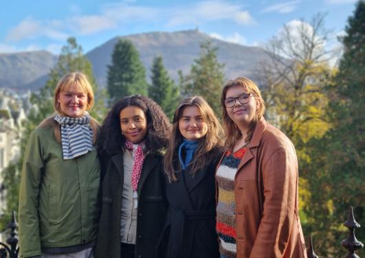Fire unge kvinner med ytterklede, tre, fjell og blå himmel i bakgrunnen