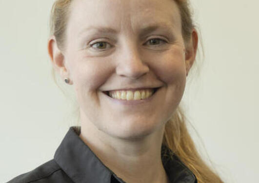 Profile picture of Silje Kristiansen, smiling and looking into the camera. Blonde long hair in a ponytail, white skin, light brown eyes, wearing a black shirt.