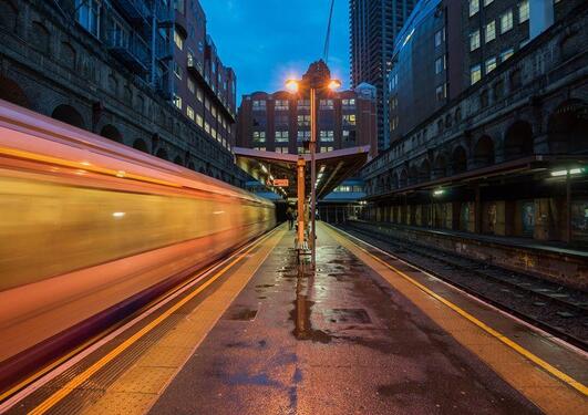 An image of a train speeding past a station in a a city-scape during dusk