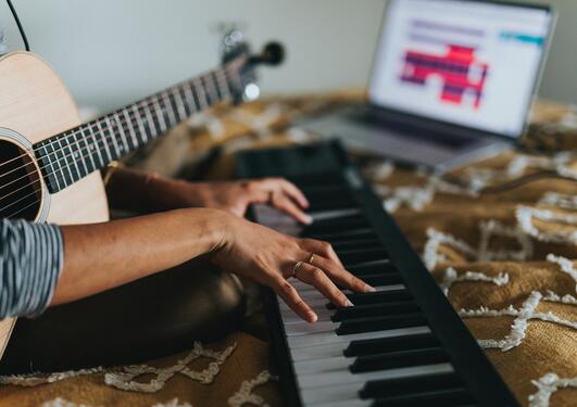 [ID] Bildet viser en person med en guitar i fanget som spiller på et piano. Et brunt teppe er under pianoet og en PC i bakgrunnen.