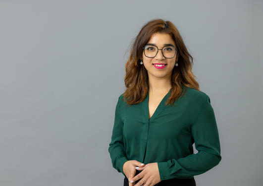 Subina Shresta is looking to the camera, smiling. She has caramel brown hair, glasses, wearing a green blouse. Her hands ar collected in front of her. The background is light grey.