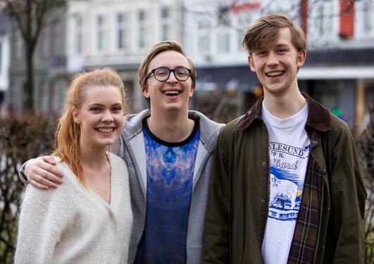 Tre engasjerte studenter smiler mot kamera med hus og park i bakgrunnen.