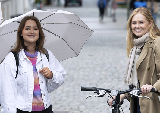 Studenter ute i gaten med paraplyer og sykkel. Tre jenter som smiler mot fotografen