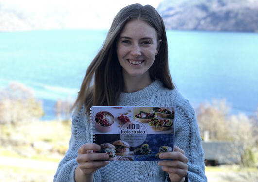 Tonje Eiane Aarsland med kokeboken sin i hånden, hun smiler. Bak henne ser man blått hav 