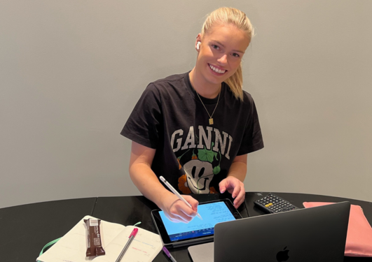 Blond jente smiler til kamera. Det er bøker og en mac foran henne på bordet.