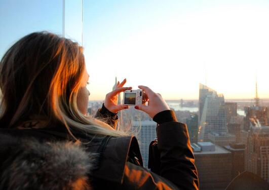 En jente ser ned på en storby fra et høyt tårn og tar bilde av solnedgangen