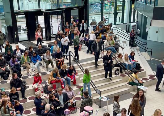 The entrance to MCB is filled with many students standing and sitting around on chairs. They are at an media event of some sort. .