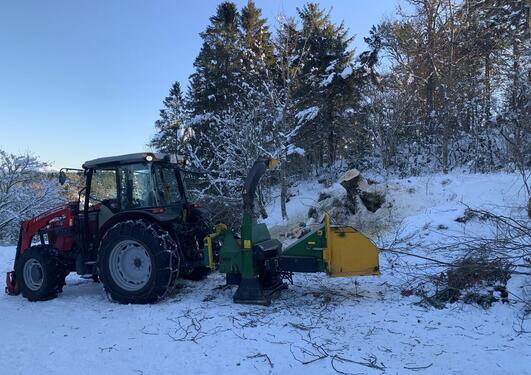En traktor med vedkutter, tre og blå himmel i bakgrunnen, snø på bakken