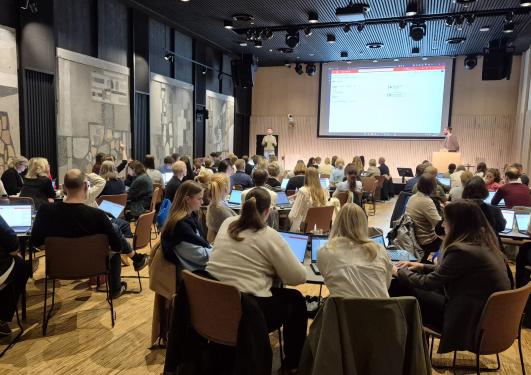 Ansatte som sitter i grupper og jobber i Storsalen, person på scenen presenterer Power Automate